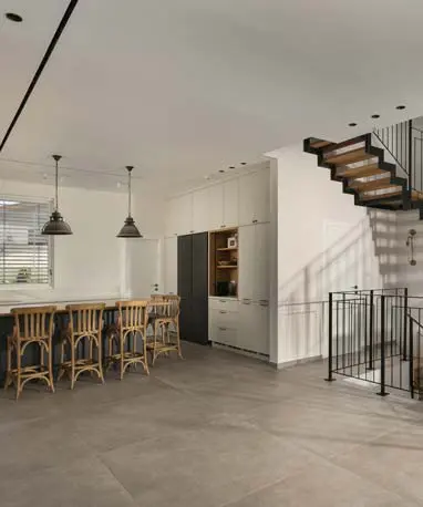 חלל ציבורי פתוח עם אי מטבח וכיסאות עץ לצד גרם מדרגות מתכת ועץ ותקרה עם מיזוג לינארי