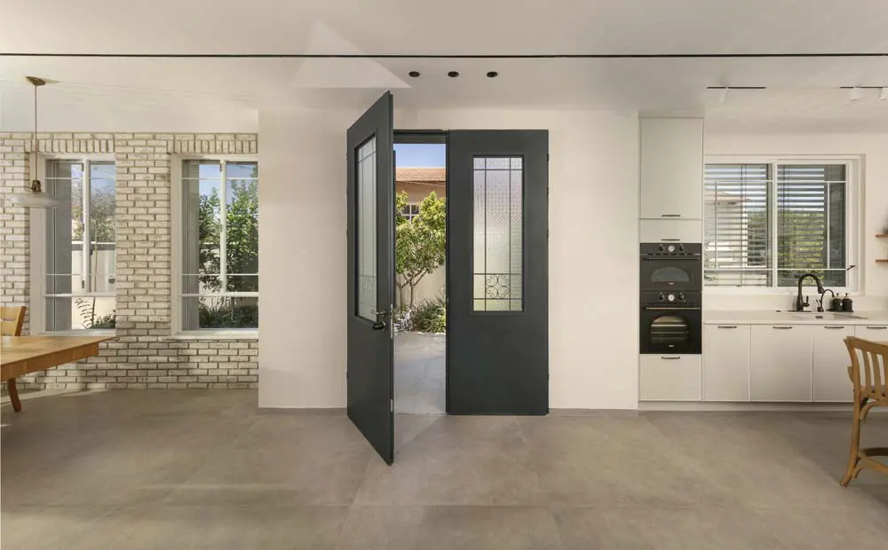 דלת כהה עם זכוכית ופרזול דקורטיבי פתוחה אל הגינה, לצד קיר בריקים ומטבח לבן